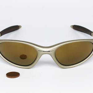 May include: A pair of Oakley sunglasses with a silver frame and brown lenses. The arms are dark brown. A penny is next to the sunglasses. The Oakley logo is visible on the frame.