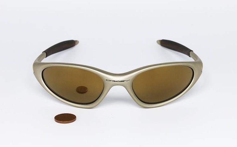 May include: A pair of Oakley sunglasses with a silver frame and brown lenses. The arms are dark brown. A penny is next to the sunglasses. The Oakley logo is visible on the frame.