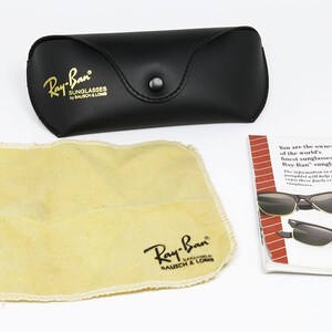 Peut inclure: Un &eacute;tui en cuir noir avec inscription dor&eacute;e "Ray-Ban Sunglasses by Bausch & Lomb". Un chiffon de nettoyage beige avec le m&ecirc;me logo est &agrave; c&ocirc;t&eacute; de l'&eacute;tui. Une brochure blanche avec le texte "You are the owner of the world's finest sunglasses, Ray-Ban sunglasses." est &eacute;galement incluse.