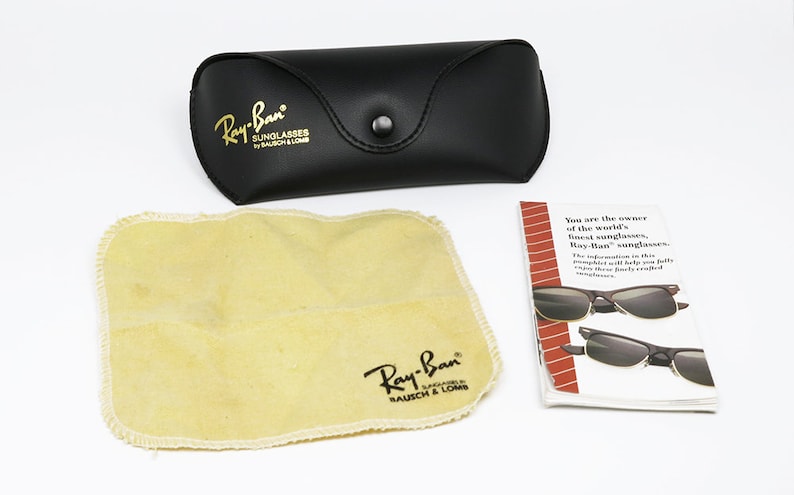 Peut inclure: Un &eacute;tui en cuir noir avec inscription dor&eacute;e "Ray-Ban Sunglasses by Bausch & Lomb". Un chiffon de nettoyage beige avec le m&ecirc;me logo est &agrave; c&ocirc;t&eacute; de l'&eacute;tui. Une brochure blanche avec le texte "You are the owner of the world's finest sunglasses, Ray-Ban sunglasses." est &eacute;galement incluse.