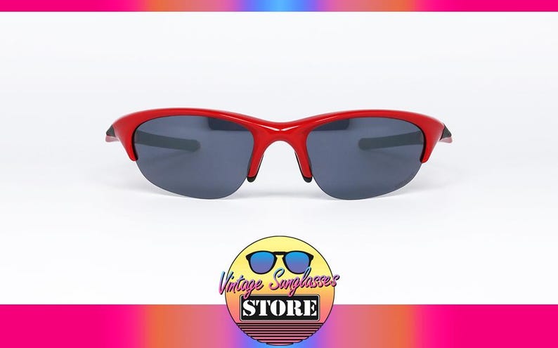 Puede incluir: Gafas de sol rojas con lentes de color gris oscuro. Las gafas tienen una montura curvada y un dise&ntilde;o deportivo. La imagen tambi&eacute;n incluye un logotipo de una tienda de gafas de sol vintage.