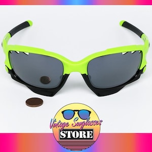 May include: Neon green and black sunglasses with gray lenses. The sunglasses have a unique design with a jagged edge on the top of the frame. A brown coin and a logo for "Vintage Sunglasses Store" are also in the image.