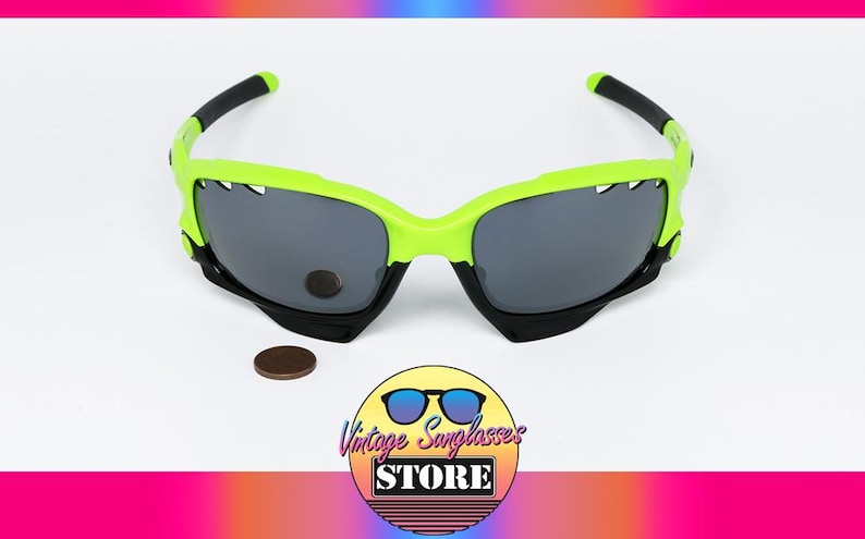 May include: Neon green and black sunglasses with gray lenses. The sunglasses have a unique design with a jagged edge on the top of the frame. A brown coin and a logo for "Vintage Sunglasses Store" are also in the image.