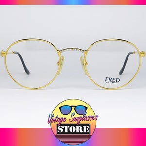 Op de afbeelding: Goudkleurige bril met een rond montuur en zwarte pootjes. De glazen zijn helder en de merknaam "FRED Paris" is op één glas gedrukt. De bril staat tegen een witte achtergrond.
