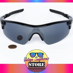 May include: Black sunglasses with gray lenses. The sunglasses have a curved design and a black frame. A penny is next to the sunglasses. The logo for Vintage Sunglasses Store is in the bottom right corner.