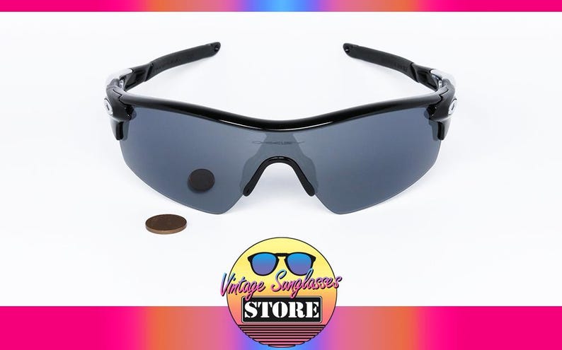 May include: Black sunglasses with gray lenses. The sunglasses have a curved design and a black frame. A penny is next to the sunglasses. The logo for Vintage Sunglasses Store is in the bottom right corner.