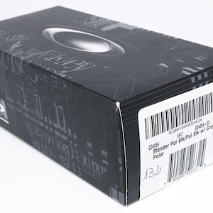 Puede incluir: Una caja rectangular negra con un dise&ntilde;o ovalado plateado en la parte superior. La caja tiene una etiqueta blanca con un c&oacute;digo de barras y el texto "Blender Pol Blk/Pol Blk w/ Grey". La caja tiene un patr&oacute;n gris oscuro.