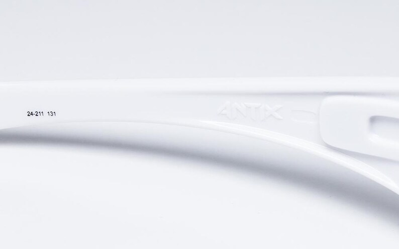 Op de afbeelding: Close-up van een wit plastic object met de tekst "ANTX" en de nummers "24-211 131" erop gedrukt. Het object heeft een gebogen vorm met een rechthoekige opening aan de rechterkant. De achtergrond is effen wit.