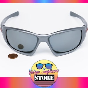 May include: Gray sunglasses with gray lenses. The sunglasses have a curved frame and a small red accent on the side. A penny is next to the sunglasses. The logo for Vintage Sunglasses Store is in the bottom corner.