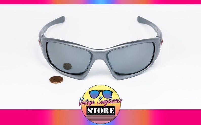 May include: Gray sunglasses with gray lenses. The sunglasses have a curved frame and a small red accent on the side. A penny is next to the sunglasses. The logo for Vintage Sunglasses Store is in the bottom corner.