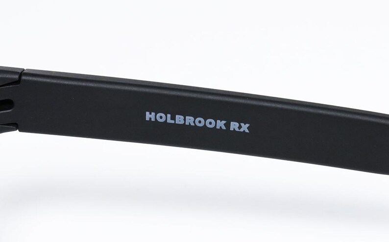 May include: A close-up of a black eyeglass arm with the text "HOLBROOK RX" in white. The arm has a matte black finish and the text is centrally positioned. The material appears to be plastic or a similar substance.