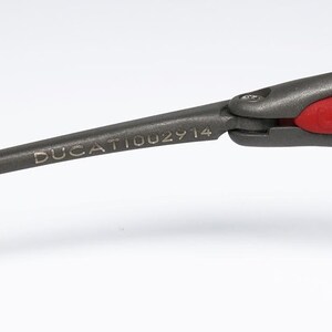 May include: Close-up of a dark gray eyeglass arm with the word "DUCATI002914" printed on it. The arm is connected to a gray frame with a red accent. The glasses are likely for sports or fashion.