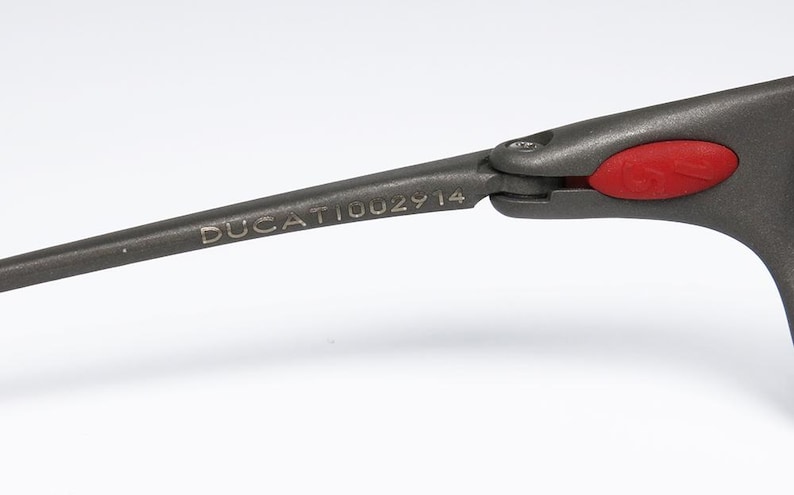 May include: Close-up of a dark gray eyeglass arm with the word "DUCATI002914" printed on it. The arm is connected to a gray frame with a red accent. The glasses are likely for sports or fashion.