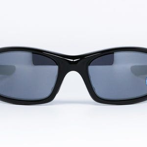 May include: Black sunglasses with a glossy finish and gray lenses. The sunglasses have a curved frame and a small blue and white sticker on the right lens. The glasses are set against a white background.