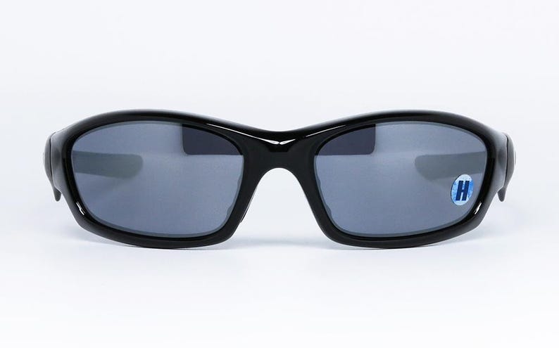 May include: Black sunglasses with a glossy finish and gray lenses. The sunglasses have a curved frame and a small blue and white sticker on the right lens. The glasses are set against a white background.