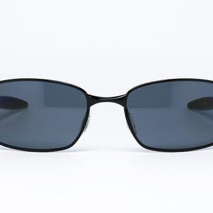 Puede incluir: Gafas de sol negras con lentes grises oscuras. Las monturas tienen un dise&ntilde;o elegante y minimalista. Las gafas de sol est&aacute;n sobre un fondo blanco, resaltando su forma y estilo. Un accesorio cl&aacute;sico para la protecci&oacute;n ocular.