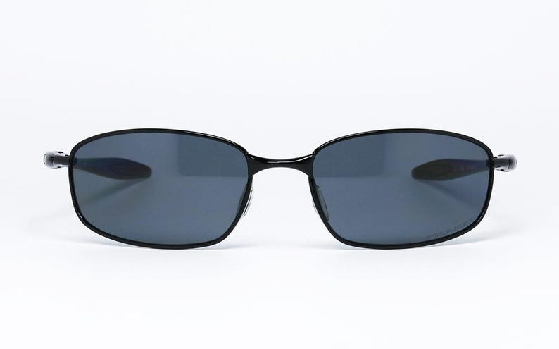 Puede incluir: Gafas de sol negras con lentes grises oscuras. Las monturas tienen un dise&ntilde;o elegante y minimalista. Las gafas de sol est&aacute;n sobre un fondo blanco, resaltando su forma y estilo. Un accesorio cl&aacute;sico para la protecci&oacute;n ocular.