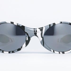 May include: Oval-shaped sunglasses with a black, white, and gray camouflage pattern on the frames. The lenses are a dark gray color, providing a sleek and modern look. These sunglasses are designed for eye protection and fashion.