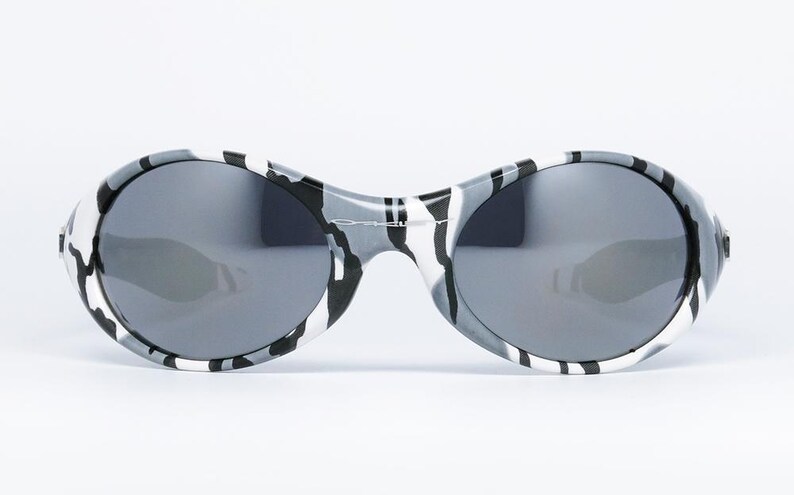 May include: Oval-shaped sunglasses with a black, white, and gray camouflage pattern on the frames. The lenses are a dark gray color, providing a sleek and modern look. These sunglasses are designed for eye protection and fashion.
