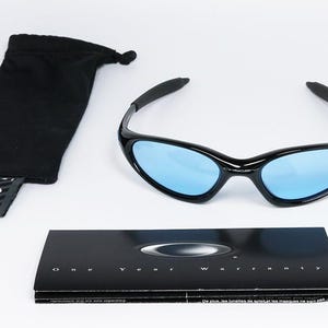 May include: Black sunglasses with blue lenses, a black drawstring pouch, and a warranty card. The sunglasses have a sleek, curved design. The text "One Year Warranty" is visible on the card. The pouch has a tag with the brand logo.