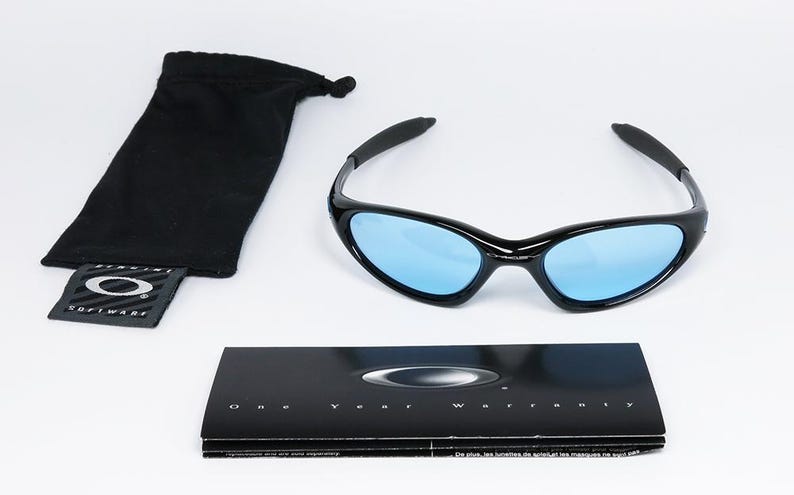 May include: Black sunglasses with blue lenses, a black drawstring pouch, and a warranty card. The sunglasses have a sleek, curved design. The text "One Year Warranty" is visible on the card. The pouch has a tag with the brand logo.