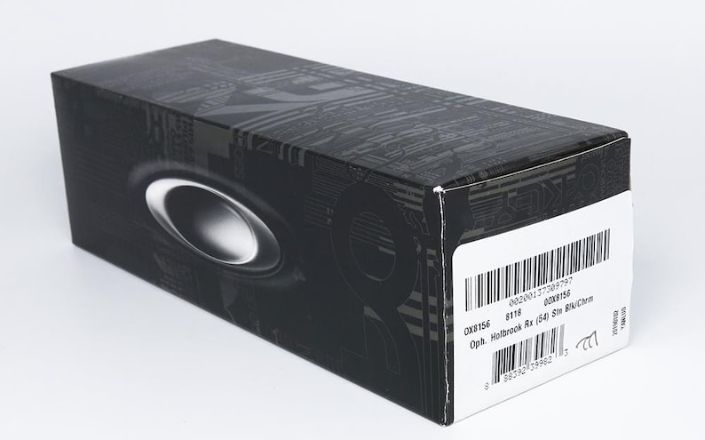 May include: Black rectangular box with a silver oval design and a white label with a barcode. The box has a textured pattern and the brand name is visible. The box is for eyewear.
