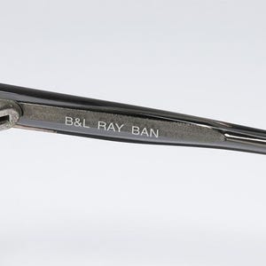 May include: Close-up of a pair of sunglasses with a dark gray frame. The text "B&L RAY BAN" is printed on the temple arm. The hinge and arm are silver-colored.