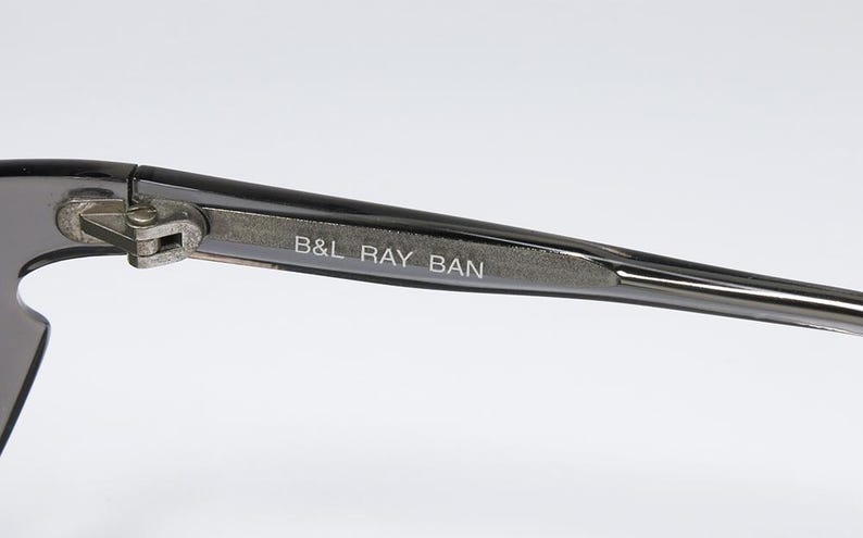 May include: Close-up of a pair of sunglasses with a dark gray frame. The text "B&L RAY BAN" is printed on the temple arm. The hinge and arm are silver-colored.
