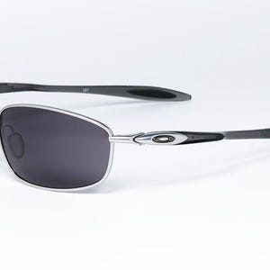 May include: Silver-framed sunglasses with dark gray lenses. The sunglasses have a sleek, modern design with black temples and a subtle logo detail. The lenses are a dark gray color, providing a contrast to the silver frame.