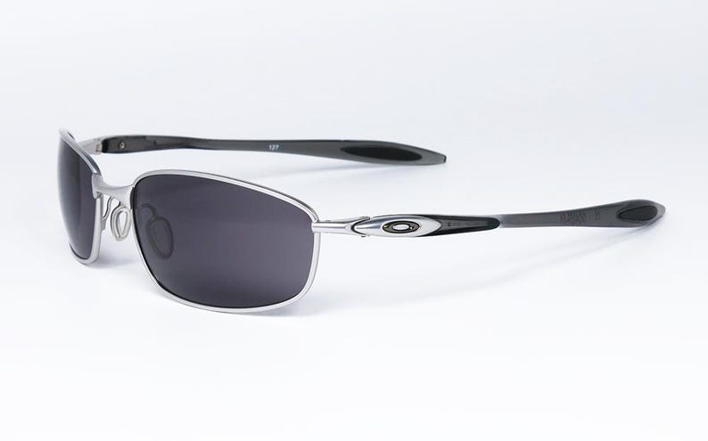 May include: Silver-framed sunglasses with dark gray lenses. The sunglasses have a sleek, modern design with black temples and a subtle logo detail. The lenses are a dark gray color, providing a contrast to the silver frame.