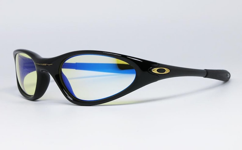 Peut inclure: Lunettes de soleil de sport noires avec une monture incurv&eacute;e et des verres teint&eacute;s bleus. Le logo Oakley est visible sur les branches et la monture. Les verres ont une teinte jaune. Con&ccedil;ues pour le sport et les activit&eacute;s de plein air.