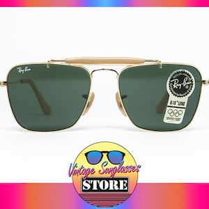 May include: Gold-framed Ray-Ban sunglasses with green lenses. Features a light brown bridge and temples. Includes a Ray-Ban sticker with Olympic rings. The sunglasses are displayed against a white background.