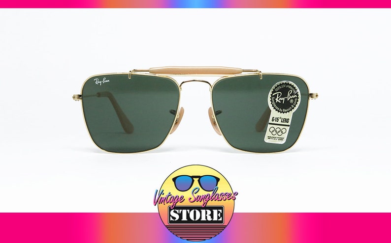 May include: Gold-framed Ray-Ban sunglasses with green lenses. Features a light brown bridge and temples. Includes a Ray-Ban sticker with Olympic rings. The sunglasses are displayed against a white background.
