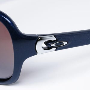 May include: Close-up of navy blue sunglasses with brown gradient lenses. The frames are a glossy navy blue with a silver hinge detail and the Oakley logo. The sunglasses are set against a white background.