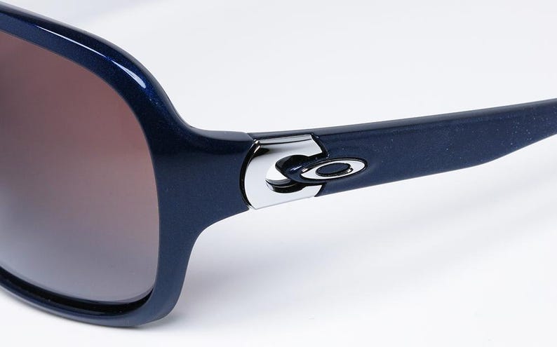 May include: Close-up of navy blue sunglasses with brown gradient lenses. The frames are a glossy navy blue with a silver hinge detail and the Oakley logo. The sunglasses are set against a white background.