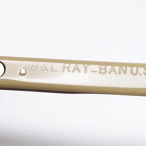 May include: Close-up of the arm of a pair of sunglasses. The arm is a light tan color and features the text "B&L RAY-BAN U.S.A." in a sans-serif font. The hinge is silver.