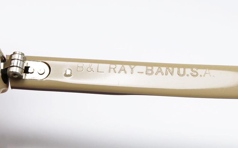 May include: Close-up of the arm of a pair of sunglasses. The arm is a light tan color and features the text "B&L RAY-BAN U.S.A." in a sans-serif font. The hinge is silver.