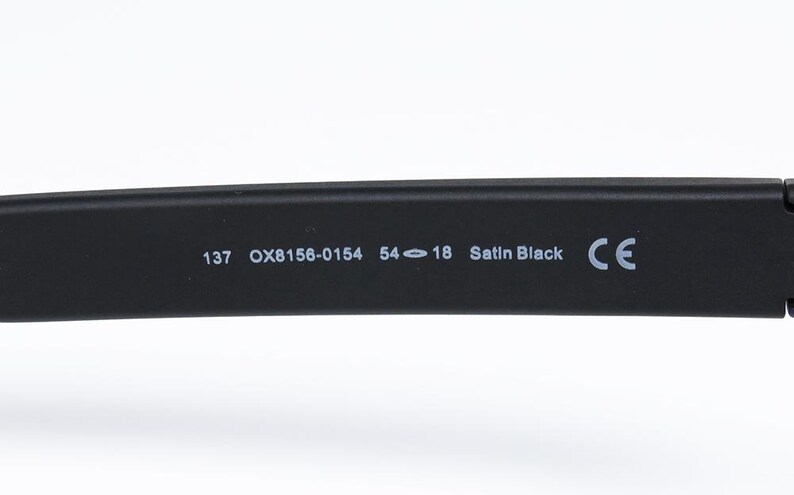 May include: Close-up of a black eyeglass arm with the following text: 137 OX8156-0154 54-18 Satin Black CE. The arm is made of a matte black material.