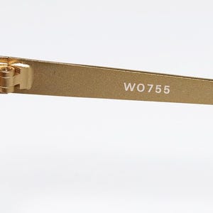 Puede incluir: Primer plano de gafas de sol doradas con lentes oscuras. La patilla de las gafas tiene el texto "WO755" en blanco. La bisagra y la montura son visibles, mostrando la construcci&oacute;n de las gafas.