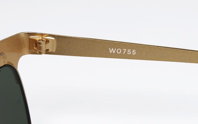 Puede incluir: Primer plano de gafas de sol doradas con lentes oscuras. La patilla de las gafas tiene el texto "WO755" en blanco. La bisagra y la montura son visibles, mostrando la construcci&oacute;n de las gafas.