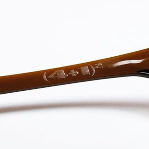 May include: A close-up shot of a curved, brown object with a shiny surface. The object has a black edge and the etched text "(4+1)&sup2;". The background is plain white.