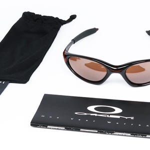 Pode incluir: &Oacute;culos de sol pretos e castanhos com uma bolsa preta com cord&atilde;o e um livreto preto. Os &oacute;culos de sol t&ecirc;m uma arma&ccedil;&atilde;o curva e lentes castanhas. O livreto apresenta o logotipo da Oakley e o texto "One Year Warranty".