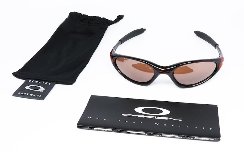 Pode incluir: &Oacute;culos de sol pretos e castanhos com uma bolsa preta com cord&atilde;o e um livreto preto. Os &oacute;culos de sol t&ecirc;m uma arma&ccedil;&atilde;o curva e lentes castanhas. O livreto apresenta o logotipo da Oakley e o texto "One Year Warranty".