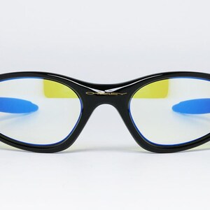 Peut inclure: Lunettes noires de forme ovale avec des branches bleues et des verres jaunes. Les lunettes ont un design &eacute;l&eacute;gant et moderne avec le nom de la marque "Oakley" en lettres dor&eacute;es sur le pont.