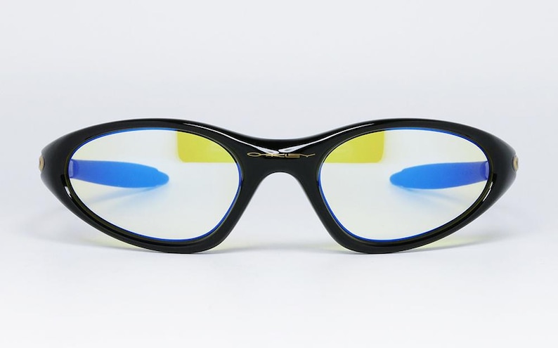 Peut inclure: Lunettes noires de forme ovale avec des branches bleues et des verres jaunes. Les lunettes ont un design &eacute;l&eacute;gant et moderne avec le nom de la marque "Oakley" en lettres dor&eacute;es sur le pont.