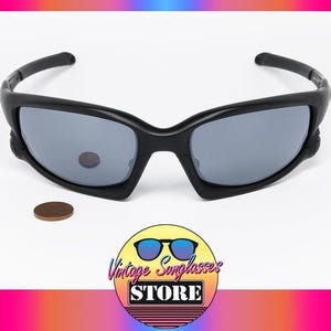 Może przedstawiać: Czarne okulary przeciwsłoneczne z szarymi soczewkami są prezentowane na białej powierzchni. Okulary przeciwsłoneczne mają sportowy design. Mały, okrągły, brązowy przedmiot znajduje się obok okularów. Obraz zawiera logo "Vintage Sunglasses Store".