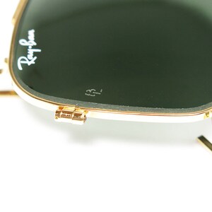 May include: Close-up of Ray-Ban sunglasses with a gold frame and dark green lenses. The Ray-Ban logo is visible in white on the upper left lens. The sunglasses are partially open, showing the hinge and arm.