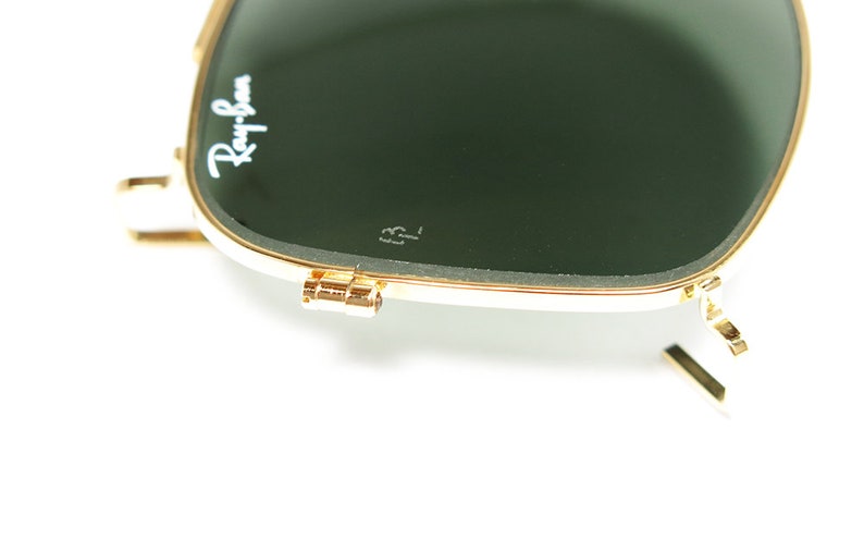 May include: Close-up of Ray-Ban sunglasses with a gold frame and dark green lenses. The Ray-Ban logo is visible in white on the upper left lens. The sunglasses are partially open, showing the hinge and arm.