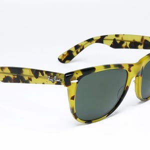 May include: Tortoiseshell-patterned sunglasses with dark green lenses. The frames are a mix of yellow and black, with the Ray-Ban logo visible on the temple. The sunglasses are angled, showcasing their design.