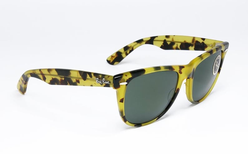 May include: Tortoiseshell-patterned sunglasses with dark green lenses. The frames are a mix of yellow and black, with the Ray-Ban logo visible on the temple. The sunglasses are angled, showcasing their design.